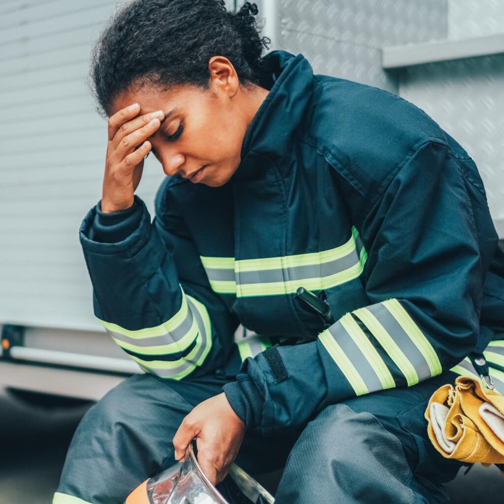 A frustrated and burned out female firefighter sits beside a fire truck holding her head.