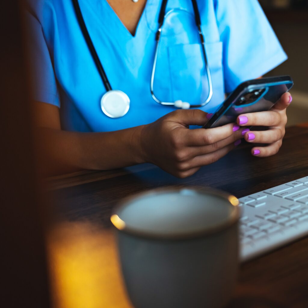 Female night shift nurse in medical office holding a smartphone and sitting in front of a computer keyboard.