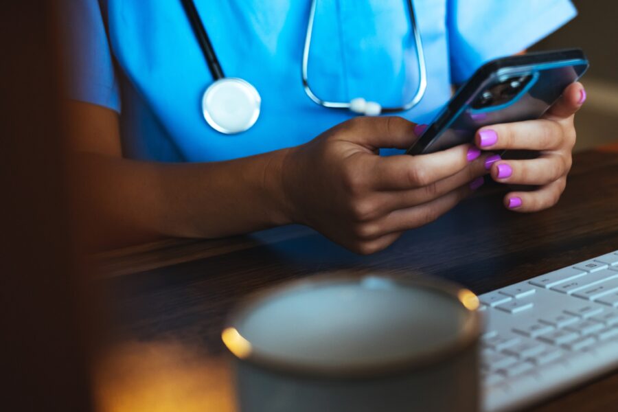 Female night shift nurse in medical office holding a smartphone and sitting in front of a computer keyboard.