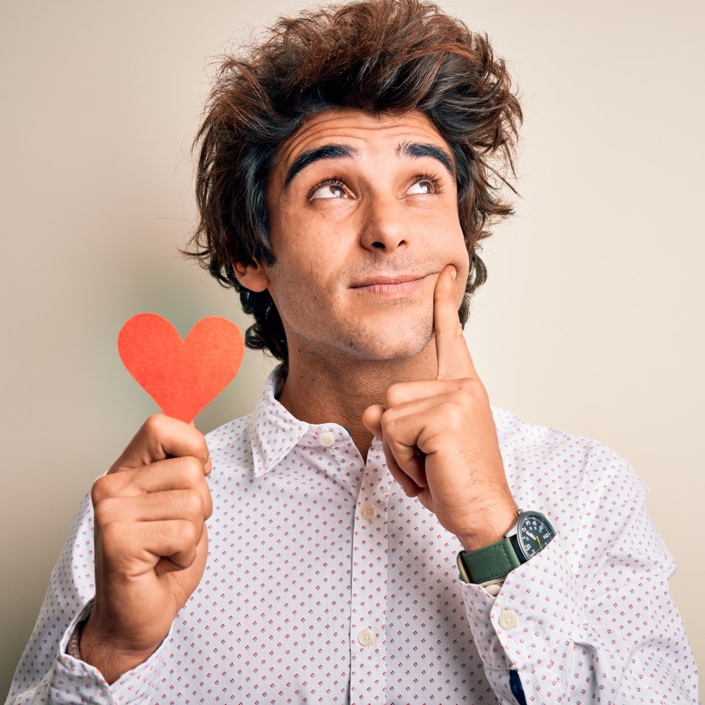 Man holding red paper heart with hand on chin thinking about Valentine's Day gifts.