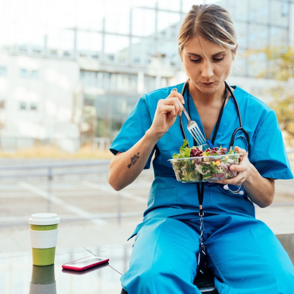 Blonde woman in blue scrubs eating a salad outside a hospital.