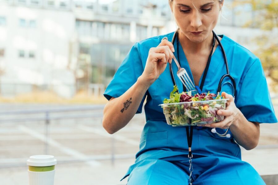 Blonde woman in blue scrubs eating a salad outside a hospital.