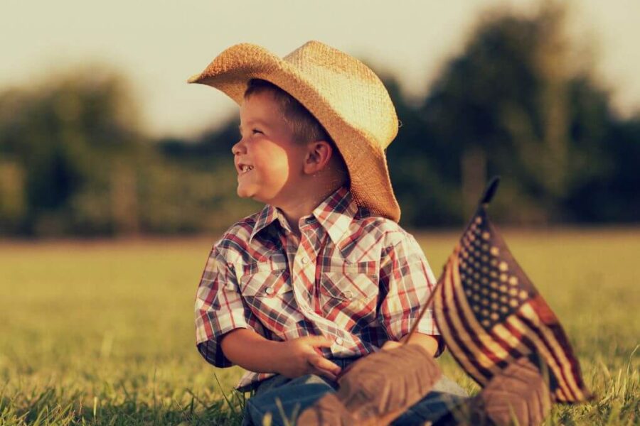 Toddler in straw cowboy hat and plaid shirt sitting in the grass holding a small U.S. flag