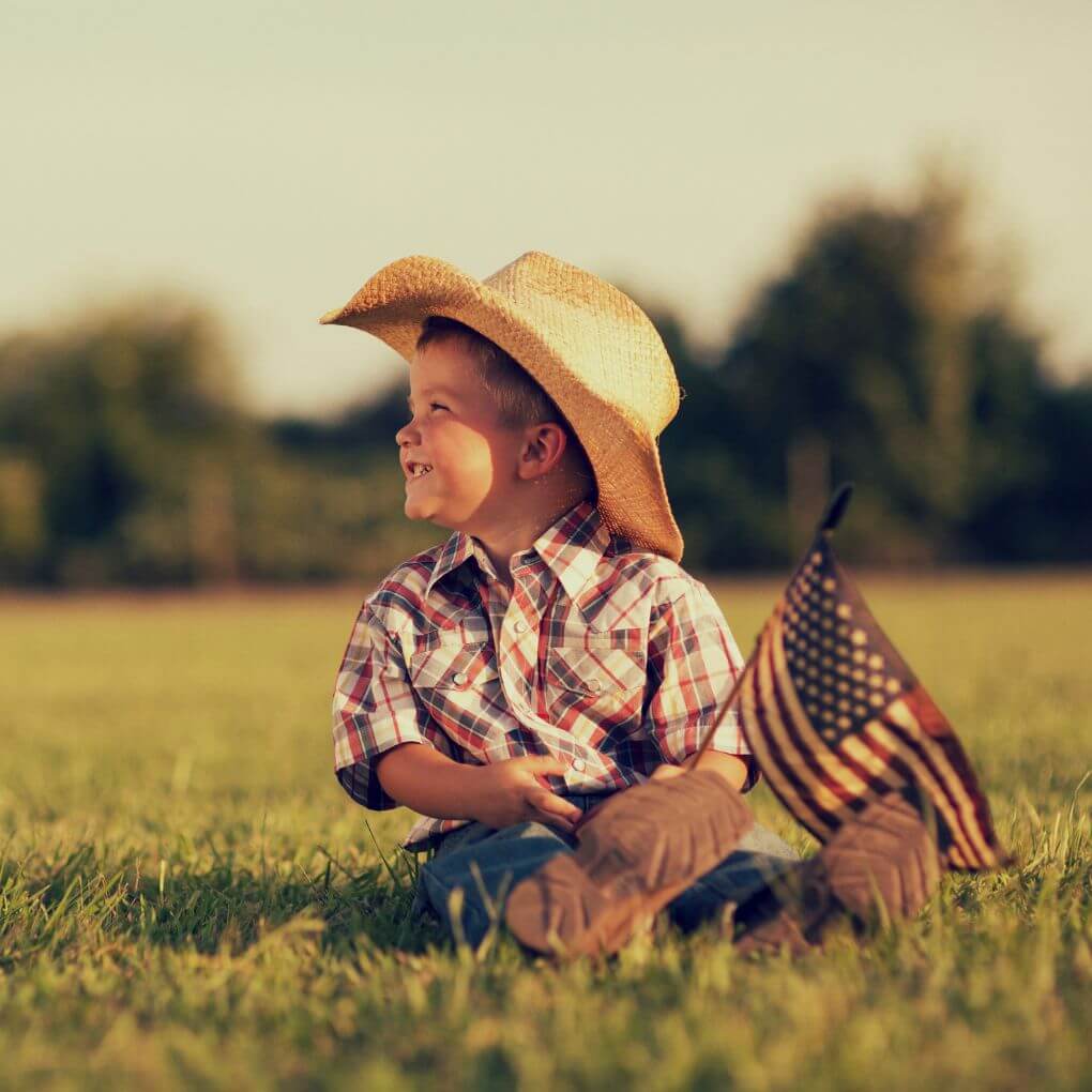 Toddler in straw cowboy hat and plaid shirt sitting in the grass holding a small U.S. flag