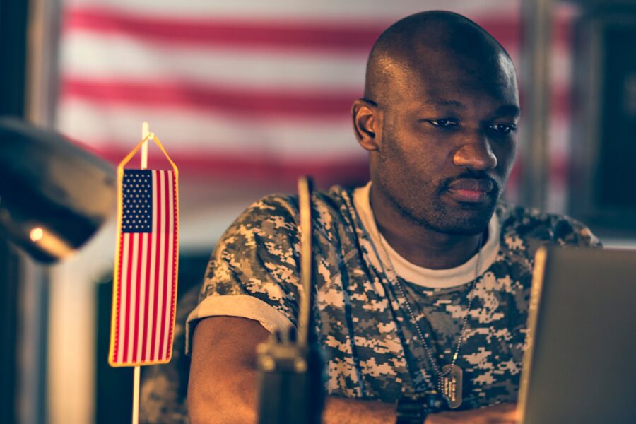 Middle-aged Black man in camo shirt using a laptop with U.S. flag in the background.