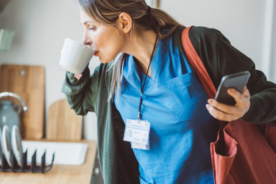 Photo of a middle-aged woman in scrubs and a jacket holding a cell phone and taking a quick sip of coffee before hurrying out the door.