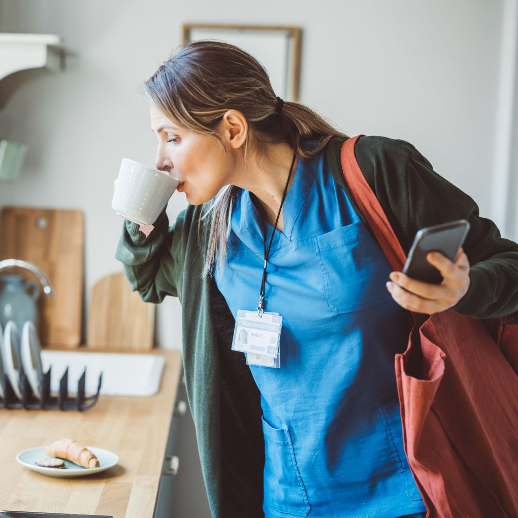 Photo of a middle-aged woman in scrubs and a jacket holding a cell phone and taking a quick sip of coffee before hurrying out the door.