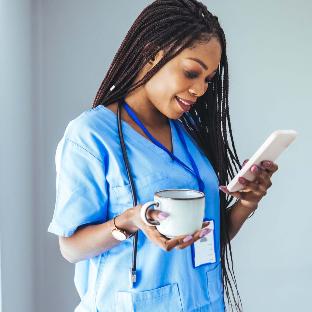 Young Black nurse in scrubs holding a coffee mug and a cell phone while ordering with her YETI nurse discount.