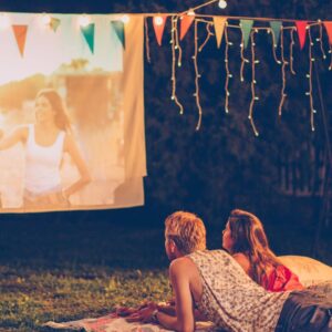 Young couple lying in their backyard on a blanket in summer watching a movie on an outdoor screen.