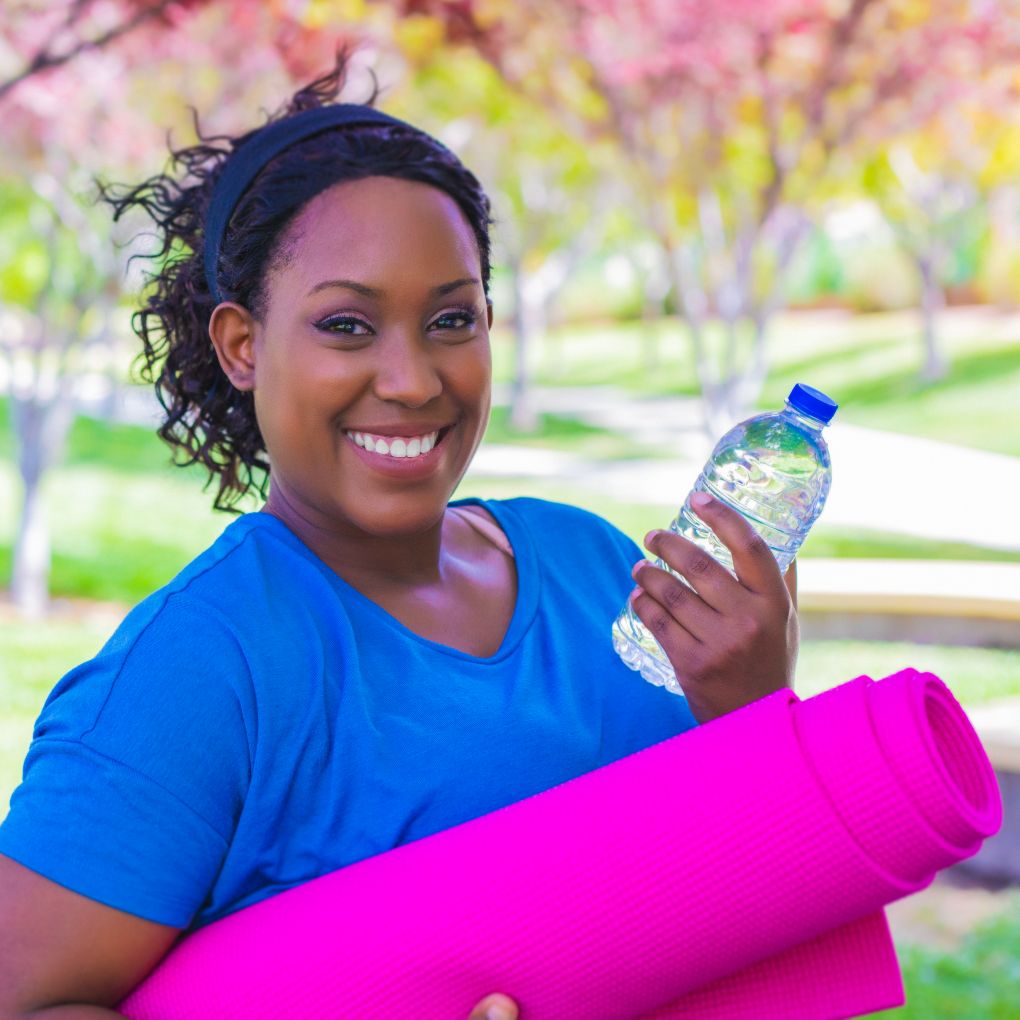 A smiling Black nurse in a blue t-shirt holding a bright pink yoga mat and water bottle.