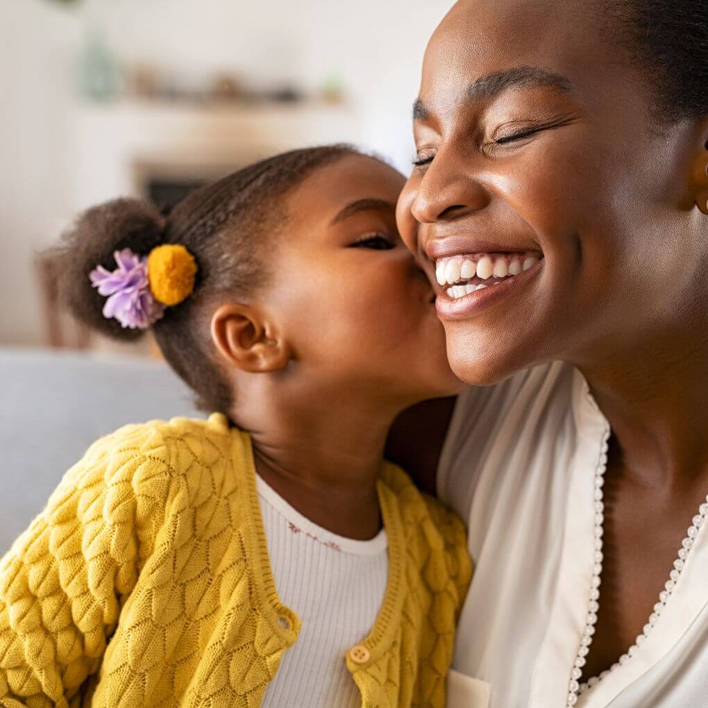 Cute Black toddler in pigtails giving her smiling mom a kiss on the cheek.