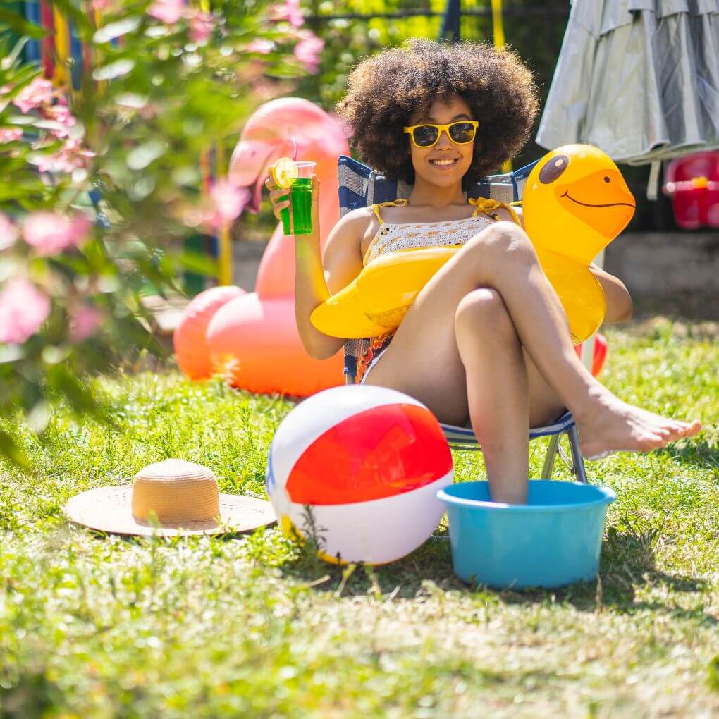 Young woman in her backyard surrounded by pool toys and sipping a drink enjoying her staycation.