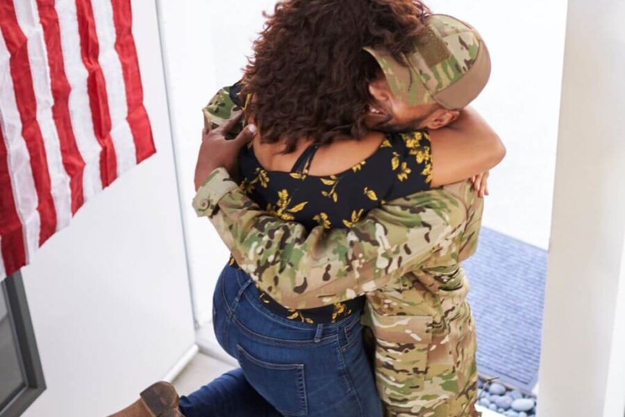 Soldier coming through the front door returning home from deployment is greeted by his spouse, who is jumping into his arms.