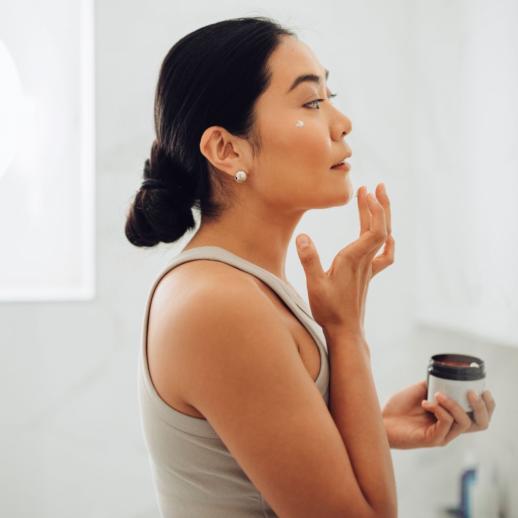 Beautiful and cheerful Asian woman standing in her bathroom, holding face cream and applying it to her cheeks and face.