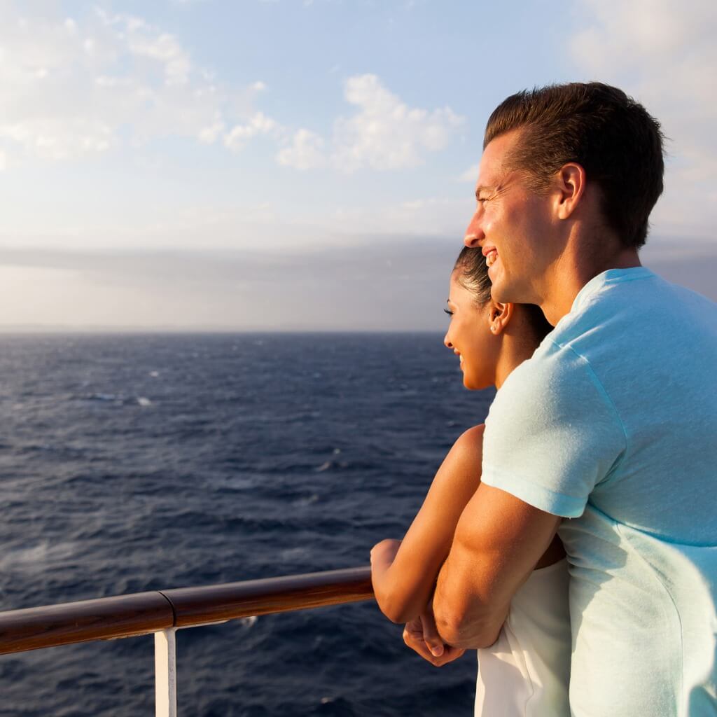 A couple embracing by a cruise ship railing with the ocean in the background, taking a budget cruise.