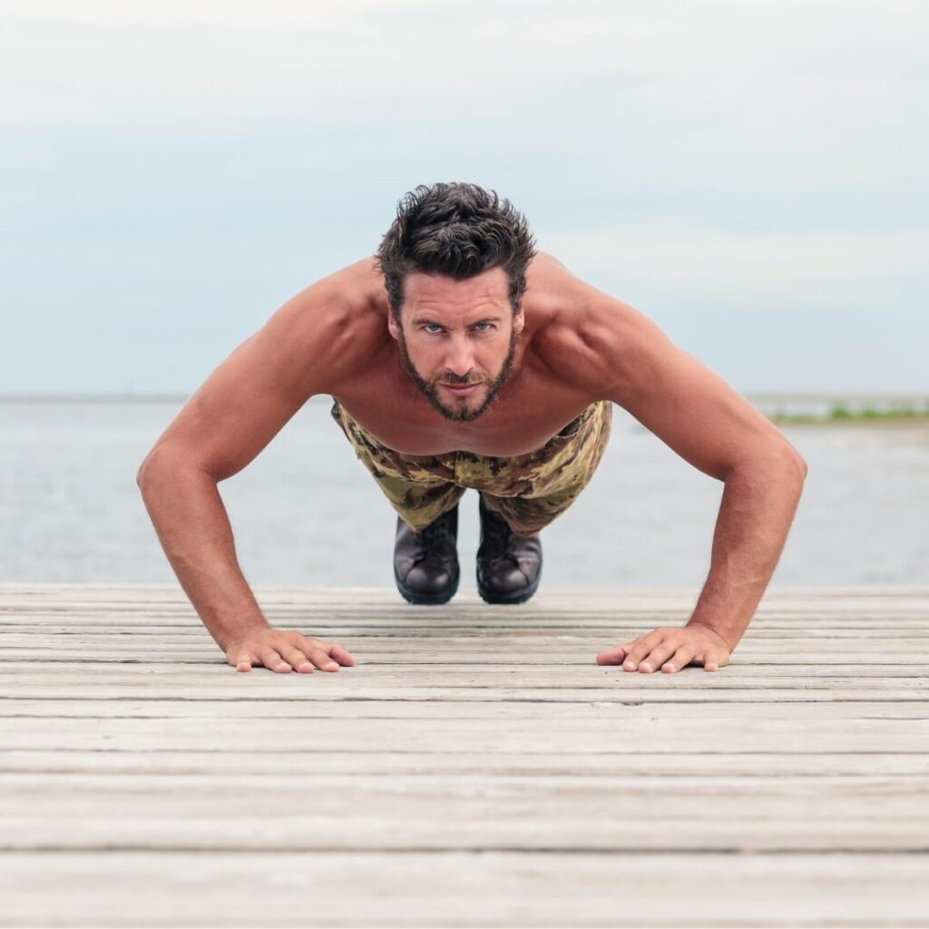 Shirtless man in camo pants and boots doing a pushup on a beach.
