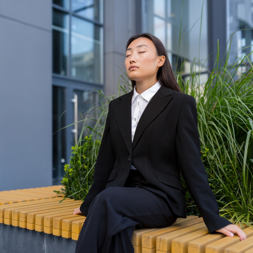 Young Asian woman in work attire meditating outside on a bench getting instant stress relief.