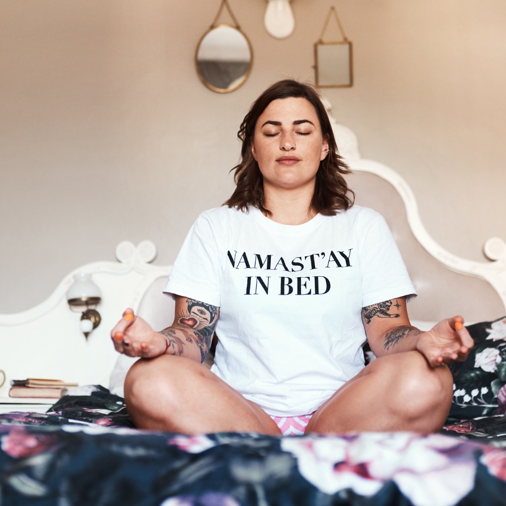 A brown-haired woman in a "Namastay in bed" t-shirt sitting in a yoga pose on her bed learning how to meditate in bed.