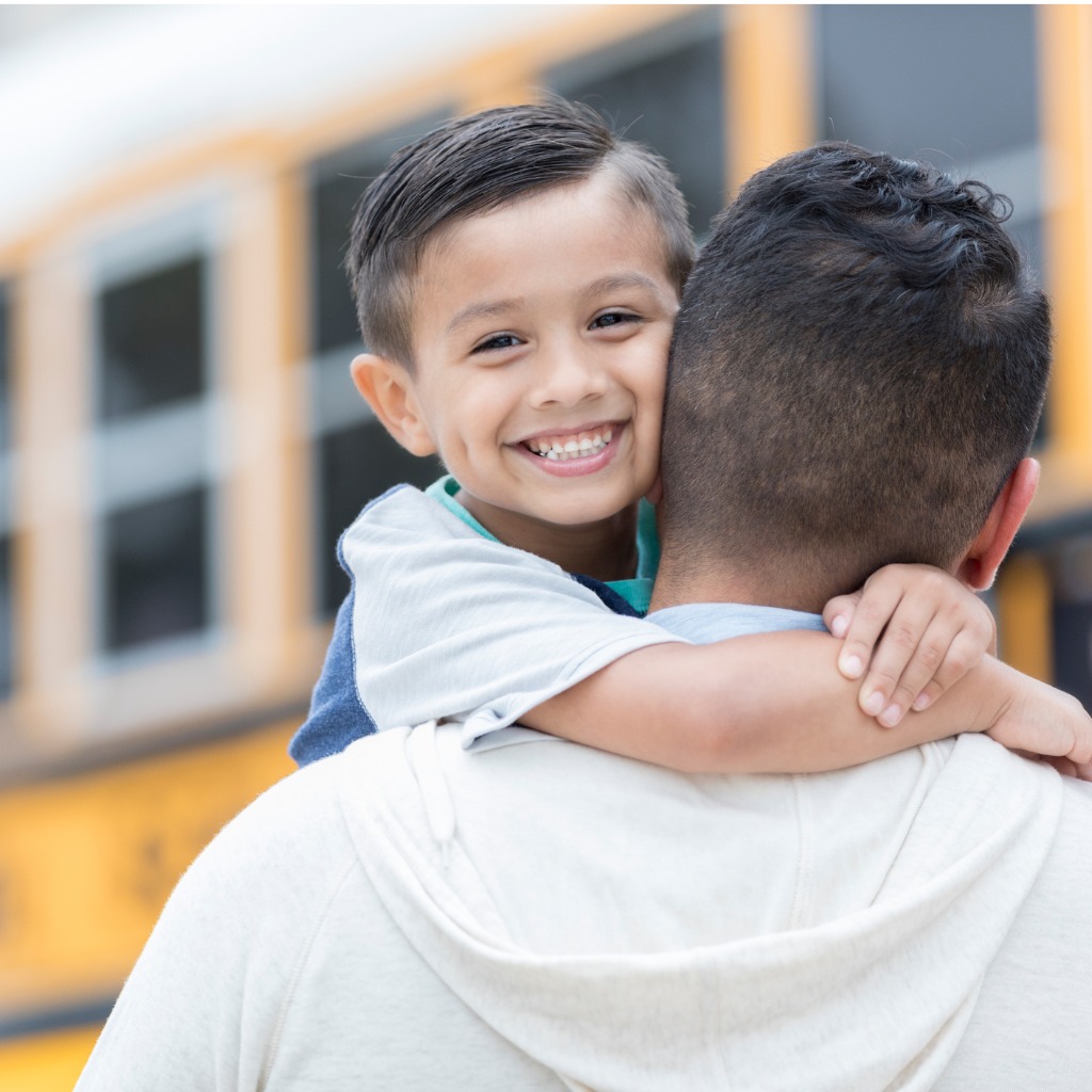 Photo of a child hugging his dad as they head toward a school bus, relevant to the Military Interstate Compact.