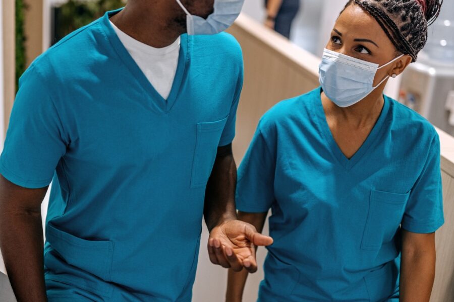 A Black male nurse talking to a Black female nurse in a hospital hallway as if telling a nursing confession.