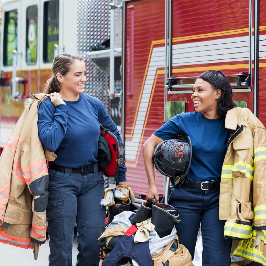 Two female firefighters smiling, holding their gear and walking away from a firetruck.