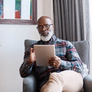 A black man with a silver beard wearing a plaid shirt and sitting in an armchair using an Apple iPad.