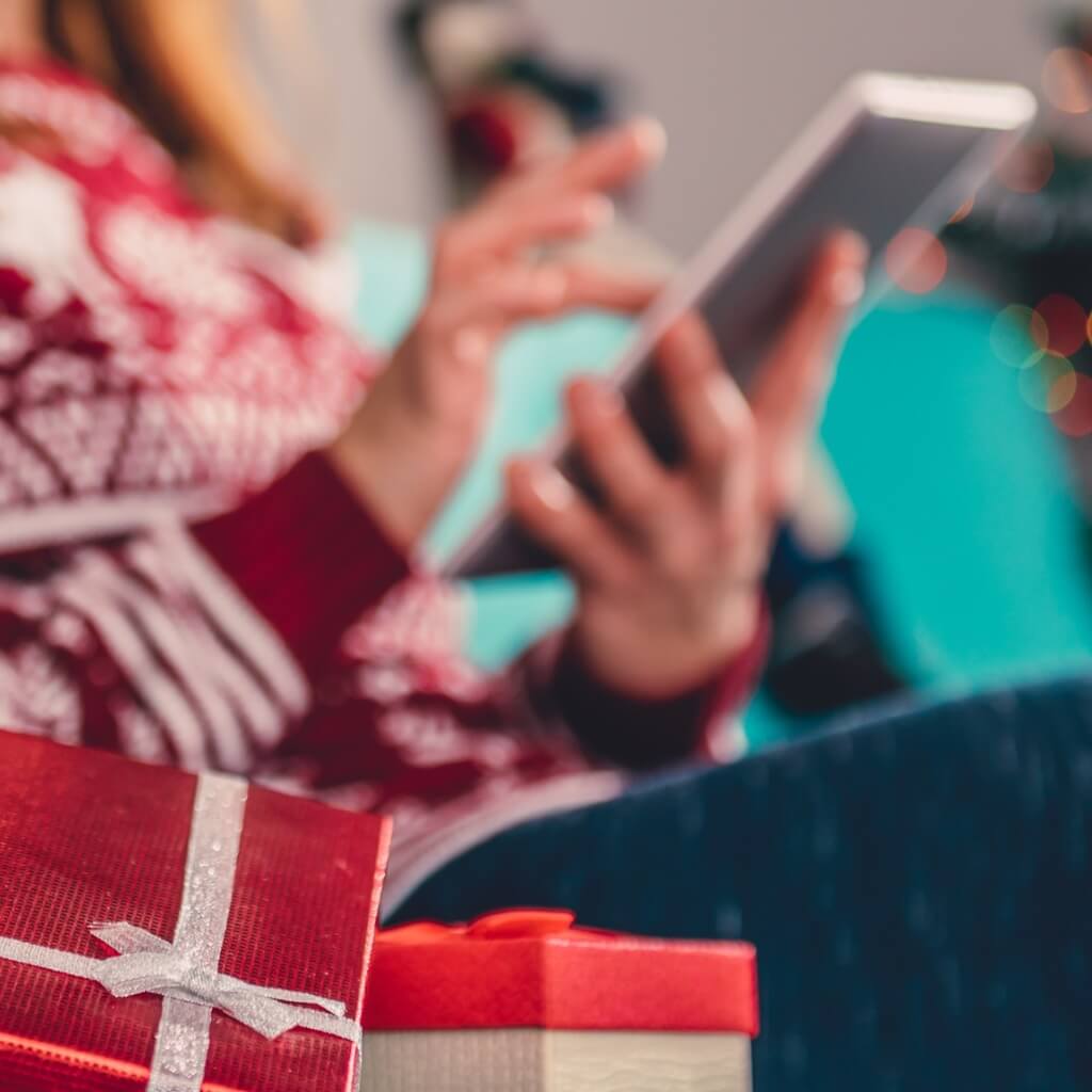 A woman in a holiday sweater holding a tablet and sitting beside wrapped presents.