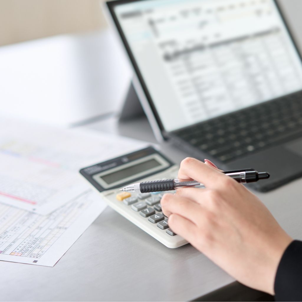 Photo of a woman's hand holding a pen and working a calculator, with tax forms and a laptop in the background.