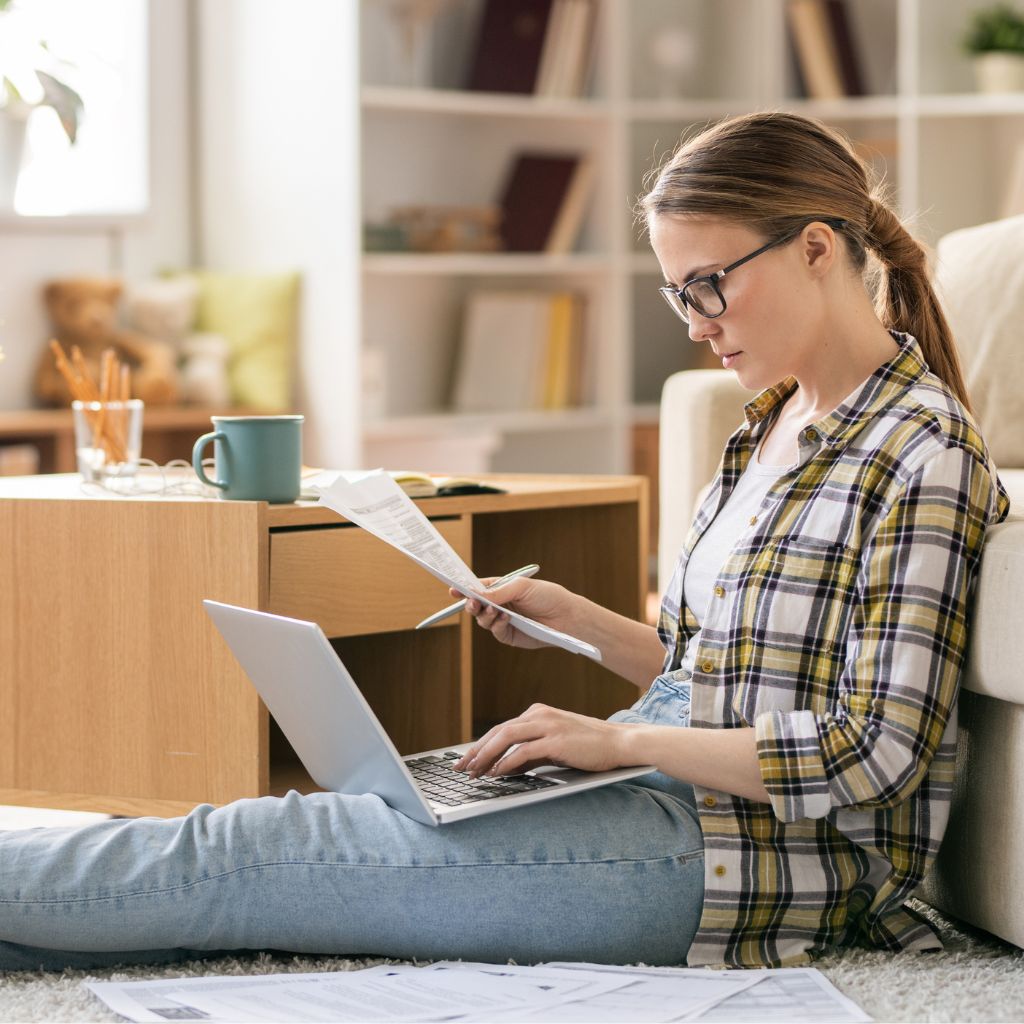 Young woman in a plaid shirt and jeans sitting on the floor with a laptop and tax papers, looking for student tax benefits.