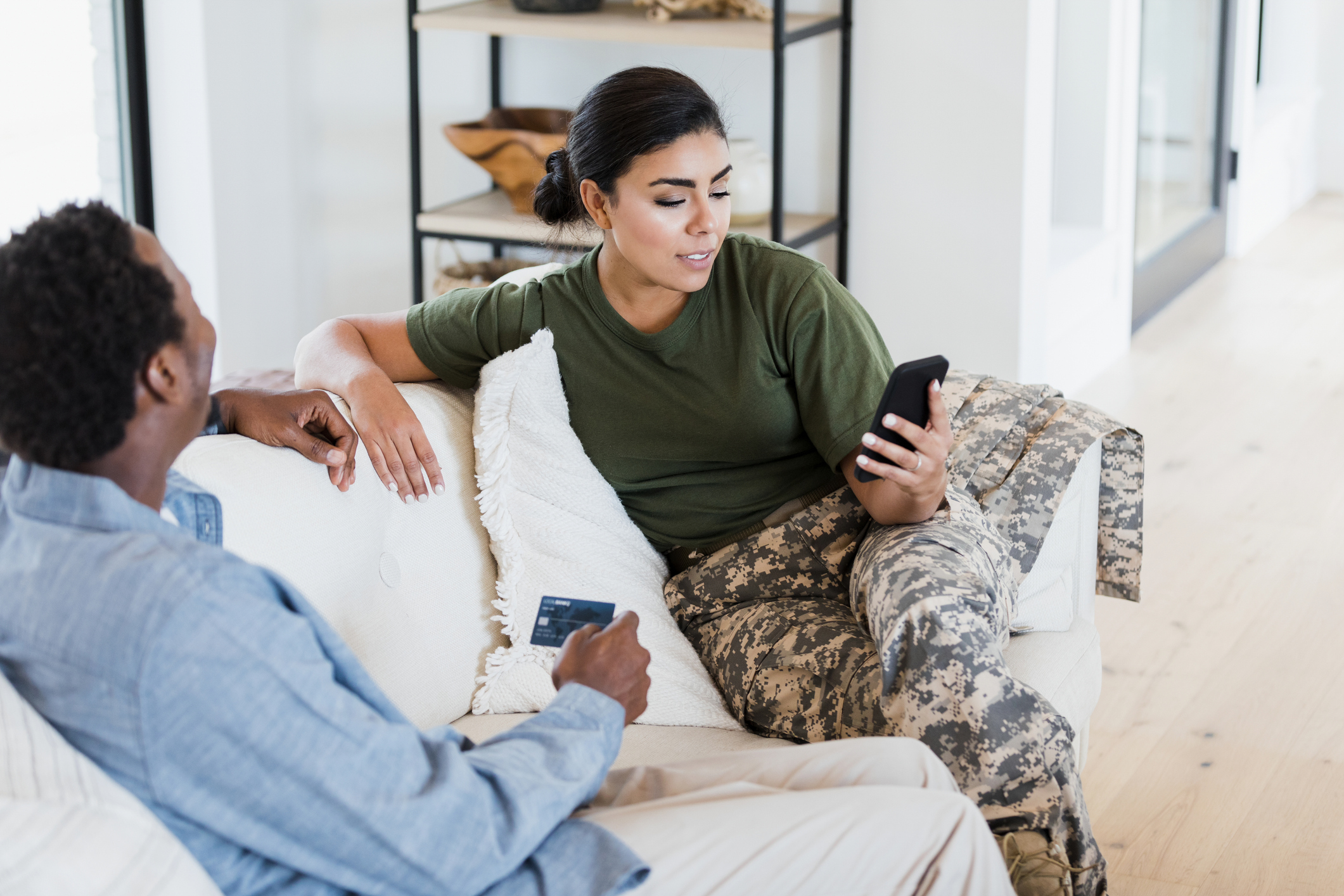 A mid adult female soldier uses a smartphone to make an online purchase. Her husband is holding a credit card.